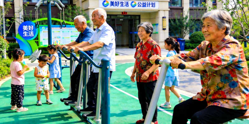 社区居民在新建的社区体育公园内进行锻炼，有老年人在使用健身器材，儿童在玩耍，背景有‘美好生活’公益计划的标识